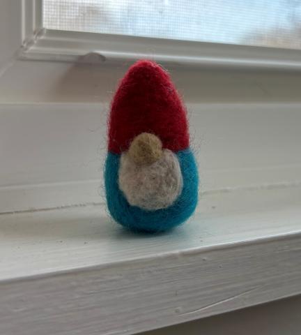 Image of a felt gnome. The gnome has a red hat and a blue body with a white beard. 