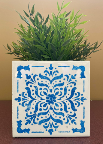 White, square, ceramic pot with blue stencil design holding a green plant. 