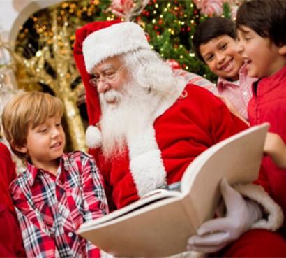 Santa reading a book to a boy in a plaid shirt with two boys next to left arm. 