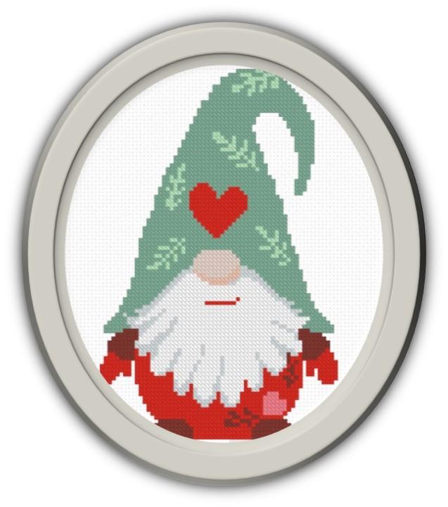 Cross stitch gnome in grey oval frame. Gnome has a sage green hat with red heart and a red outfit.